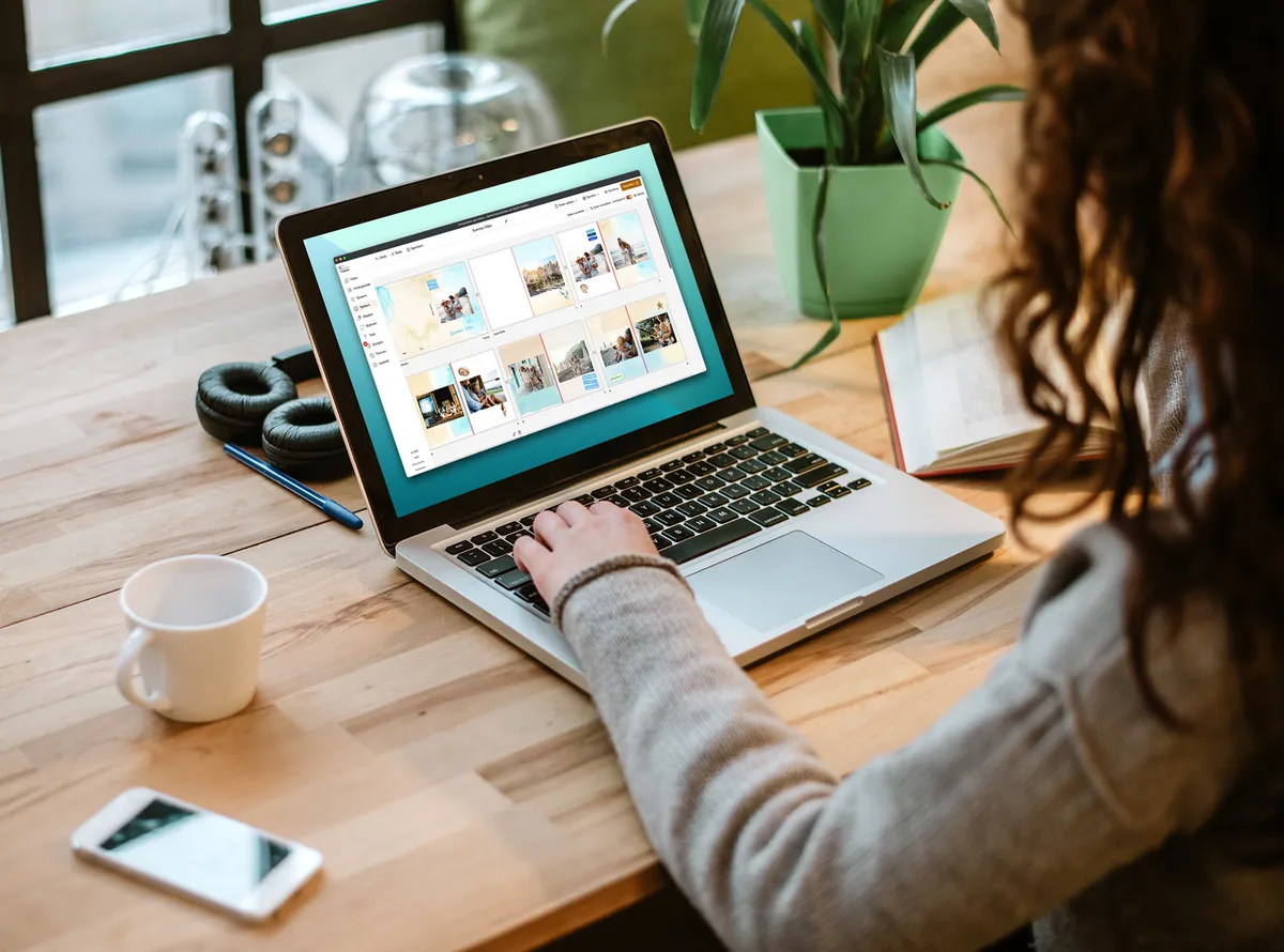 Frau sitzt einem Holztisch und hat einen Laptop vor sich, an dem sie in der PRO1 Desktop App im Fotoprodukt-Designer arbeitet und ein Fotobuch gestaltet